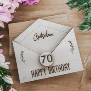personalisierter Holz-Gutschein Umschlag mit Gutschein mit Gravur „Gutschein“, „70“ und „Happy Birthday“, verziert mit floralen Elementen, auf einem Holztisch mit Blumen und Deko.