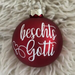Rote matte Weihnachtskugel mit weissem Schriftzug „beschts Gotti“, fotografiert auf weichem, cremefarbenem Fellhintergrund.