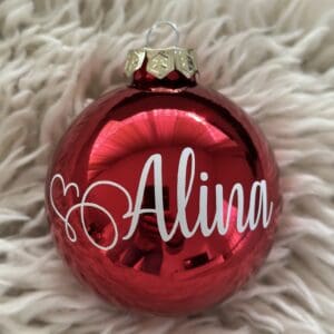 Glänzende kirschrote Weihnachtskugel mit weissem Namensschriftzug „Alina“, dekorativ fotografiert auf weichem cremefarbenem Fell.