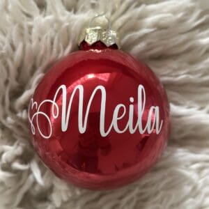 Glänzende rote Weihnachtskugel mit weissem Namen „Meila“, fotografiert auf weichem cremefarbenem Fellhintergrund.