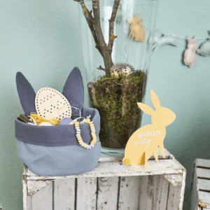 Blaues Osternest aus Stoff mit Hasenohren, gefüllt mit Holzspielzeug und Perlenkette, dekorativ inszeniert auf Kiste