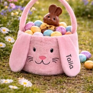 Personalisierter Osterhasenkorb aus rosafarbenem Plüsch, gefüllt mit Ostereiern und Schokohase, im Frühlingsgarten. Dieses Kinder Osternest mit Hasenohren personalisiert wird individuell mit Name beschriftet und ist das perfekte Ostergeschenk für Kinder.